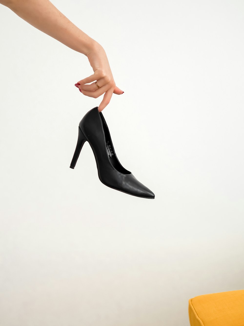 Eleganter schwarzer High Heel, an einem Finger vor weißer Wand gehalten